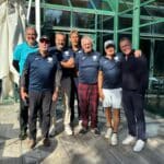 Groupe d'hommes souriants posant ensemble à l'extérieur d'un bâtiment en verre, probablement après une activité sportive ou un événement. Tous portent des tenues de sport avec un logo, ce qui suggère un lien avec une équipe ou un club. L'arrière-plan montre une architecture moderne et lumineuse, avec des éléments naturels visibles. Cette image pourrait être utilisée pour illustrer des événements sportifs ou des rencontres amicales dans un cadre convivial.