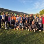 Groupe de golfeurs réunis sur un parcours, posant ensemble sous un ciel dégagé. L'arrière-plan présente des collines verdoyantes et des arbres aux couleurs d'automne. Les participants, d'âges variés, portent des vêtements de golf adaptés à la saison, et affichent des sourires, témoignant d'une ambiance conviviale. Cette image illustre un événement de golf communautaire, mettant en avant la camaraderie et le plaisir du sport en plein