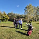 Quatre golfeurs posent sur un parcours de golf ensoleillé, entourés d'une verdure luxuriante et d'arbres aux feuilles colorées. Ils portent des vêtements de sport adaptés pour le golf et se tiennent près de leurs sacs de golf. En arrière-plan, un panneau d'information est visible, indiquant que l'endroit est un terrain de golf. Cette image illustre une journée agréable de golf entre amis, mettant en avant la convivialité et le plaisir