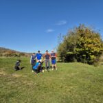 Groupe de golfeurs sur un parcours ensoleillé, entourés de verdure et d'arbres aux feuilles d'automne. Les golfeurs, équipés de chariots de golf, se préparent à jouer, affichant un esprit convivial. En arrière-plan, une colline et un drapeau de putting green sont visibles, illustrant une belle journée de golf en plein air.
