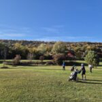 Un groupe de golfeurs sur un parcours verdoyant, entouré de collines boisées aux couleurs automnales. Le ciel est dégagé, offrant une belle lumière pour la pratique du golf. Des chariots de golf sont visibles, signalant une journée active sur le terrain. Les arbres en arrière-plan ajoutent une touche de nature, créant un cadre paisible pour les amateurs de golf. Cette image illustre parfaitement une journée agréable de jeu en plein air.