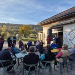 Un groupe de personnes assiste à une réunion en plein air devant le bâtiment de l'Association Sportive Golf des Prisons. Les participants, vêtus de vêtements décontractés, écoutent attentivement un discours. En arrière-plan, un paysage automnal avec des arbres aux feuilles colorées et un ciel bleu clair. Cette scène illustre la convivialité et l'engagement communautaire autour des activités sportives.