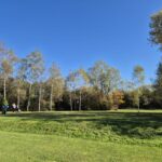 Vue d'un parcours de golf sous un ciel bleu dégagé, avec des arbres feuillus en arrière-plan. Deux golfeurs sont visibles, l'un se préparant à frapper une balle et l'autre attendant. L'herbe est bien entretenue, créant un cadre idéal pour la pratique du golf. Cette image illustre une journée ensoleillée propice aux activités de plein air.