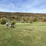 Un golfeur marche sur un parcours verdoyant, tirant un chariot de golf. En arrière-plan, des arbres aux couleurs d'automne entourent le terrain, créant une ambiance pittoresque. D'autres golfeurs profitent également de la journée ensoleillée, mettant en avant l'attrait des activités de plein air et du golf pendant la saison automnale. Ce paysage naturel offre une vue agréable et invite à la détente et au sport en harmonie avec la nature.