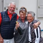 Groupe de trois personnes posant ensemble, souriantes, lors d'une célébration. L'homme à gauche porte un gilet noir et un polo rouge, tandis que la femme au centre, tenant une bouteille de champagne, porte un sweat gris. La troisième femme, à droite, est vêtue d'une veste à motifs floraux. L'arrière-plan montre un mur blanc avec des fenêtres. Cette image évoque des moments de convivialité et de joie partagée.