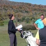 Une instructrice de golf discute avec des golfeurs sur un parcours verdoyant, entouré de collines aux feuillages automnaux. Elle tient un document dans ses mains, tandis que les participants écoutent attentivement. L'ambiance est conviviale, reflet d'une séance d'entraînement dédiée à l'amélioration des compétences en golf. Les golfeurs sont équipés de chariots et portent des vêtements adaptés à la pratique du sport en plein air.