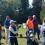 Groupe de golfeurs attentifs lors d'une séance d'instruction sur un parcours de golf. On peut voir des golfeurs de différents âges, certains portant des vêtements de sport adaptés, tandis que d'autres se tiennent à proximité de leurs chariots de golf. L'environnement est verdoyant, avec des arbres en arrière-plan, représentant une belle journée ensoleillée propice au golf. Les participants semblent engagés et intéressés par les conseils donnés.