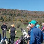 Groupe de golfeurs rassemblés sur un parcours pendant une journée ensoleillée d'automne. Une instructrice souriante donne des conseils aux participants, entourés de chariots de golf. En arrière-plan, des arbres aux feuilles colorées créent une atmosphère pittoresque. Cette image illustre une activité de golf communautaire, favorisant l'apprentissage et le partage entre passionnés.