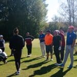 Groupe de golfeurs sur un parcours ensoleillé, assistant à une démonstration ou une séance d'instruction. Les participants portent des vêtements de sport adaptés au golf, tandis que des sacs de golf sont visibles en arrière-plan. L'environnement verdoyant souligne une belle journée de golf, idéale pour apprendre et partager des conseils entre joueurs.