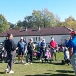 Groupe de golfeurs rassemblés sur un terrain de golf par une belle journée ensoleillée. Une instructrice se tient devant eux, expliquant des techniques ou des stratégies de jeu. Les participants, d'âges variés, écoutent attentivement, avec leurs sacs de golf à proximité. L'ambiance est conviviale et axée sur l'apprentissage du golf. En arrière-plan, on aperçoit des arbres aux couleurs d'automne et un bâtiment, ajoutant