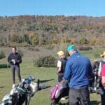 Groupe de golfeurs en formation sur un terrain de golf, avec un coach présentant des conseils. Les participants, vêtus de tenues adaptées, écoutent attentivement, entourés par un paysage automnal coloré. Des sacs de golf sont visibles au premier plan, tandis qu'un ciel bleu clair complète la scène. Cette image illustre l'importance de l'apprentissage et de la camaraderie dans le golf.