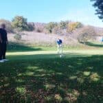 Trois golfeurs pratiquent sur un green en automne. Deux hommes observent un joueur en train de putter, tandis qu'un troisième se tient légèrement en retrait, prêt à jouer. Le paysage environnant présente des arbres aux couleurs automnales et un ciel clair. L'image évoque une ambiance conviviale et détendue, typique d'une journée de golf en plein air.