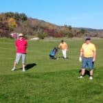 Trois golfeurs profitent d'une journée ensoleillée sur un parcours de golf. L'un d'eux se tient debout sur le green, vêtu d'un polo rouge et d'un chapeau, tandis que deux autres, habillés en orange, avancent avec un chariot de golf. En arrière-plan, on aperçoit des arbres aux couleurs d'automne et un ciel bleu clair, créant une atmosphère agréable pour la pratique du golf. Cette scène illustre la