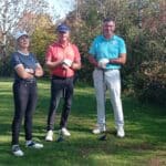 Trois golfeurs se tiennent sur un parcours verdoyant, entourés d'arbres aux feuilles d'automne. La femme à gauche porte un haut noir et un pantalon noir, tandis que l'homme au centre est vêtu d'une chemise rouge et l'autre homme à droite arbore un polo bleu. Tous tiennent un club de golf, prêts à jouer. L'image évoque une belle journée de golf, idéale pour profiter de l'extérieur et des loisirs en plein air.