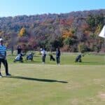 Un groupe de golfeurs sur un parcours de golf par une journée ensoleillée d'automne. L'un des joueurs se prépare à putter sur le green, tandis que d'autres attendent leur tour près de leurs chariots de golf. En arrière-plan, un paysage coloré avec des arbres aux feuilles d'automne crée une atmosphère pittoresque. Cette scène illustre une journée agréable de golf, parfaite pour les amateurs de ce sport.