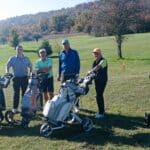 Quatre golfeurs posent sur un parcours verdoyant par une belle journée ensoleillée. Ils sont entourés de chariots de golf, prêts à profiter d'une partie. L'arrière-plan présente des collines parées de couleurs automnales, ajoutant une touche pittoresque à la scène. Cette image illustre l'esprit convivial et le plaisir du golf en plein air, parfait pour les amateurs de ce sport.