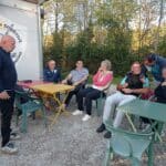 Groupe de personnes réunies autour de tables colorées à l'extérieur d'un bâtiment portant le logo de l'Association Sportive de Golf. L'ambiance semble conviviale, avec des échanges animés entre les membres. En arrière-plan, un paysage verdoyant crée un cadre agréable. Cette image illustre la vie associative et la camaraderie au sein d'un club de golf, favorisant les interactions sociales et le partage d'expériences.
