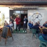 Image d'une cérémonie de remise de prix à l'Association Sportive Golf des Périsons. Des membres applaudissent alors qu'un homme en tenue sportive et une femme avec un document prennent la parole devant un groupe de personnes assises à des tables colorées. En arrière-plan, le bâtiment de l'association est visible, avec un panneau affichant le logo et des informations sur les activités. Cette scène illustre l'esprit communautaire et convivial du golf, mettant en avant l'engagement des participants