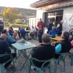 Une réunion conviviale se déroule à l'extérieur d'un bâtiment associatif, rassemblant des membres d'une association sportive. Les participants, principalement des seniors, sont assis autour de tables, attentifs aux intervenants qui s'adressent à eux. L'environnement paisible est marqué par des arbres aux feuilles d'automne en arrière-plan, créant une atmosphère chaleureuse et accueillante. Cette scène illustre l'importance de l'engagement communautaire et des activités sociales au sein