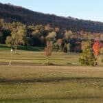 Vue panoramique d'un parcours de golf en automne, avec des arbres aux feuilles colorées en arrière-plan. Le green est bien entretenu, et un drapeau blanc indique le trou. La lumière douce du matin met en valeur le paysage vallonné, créant une atmosphère sereine et idéale pour les golfeurs. Parfait pour une journée de détente en plein air.