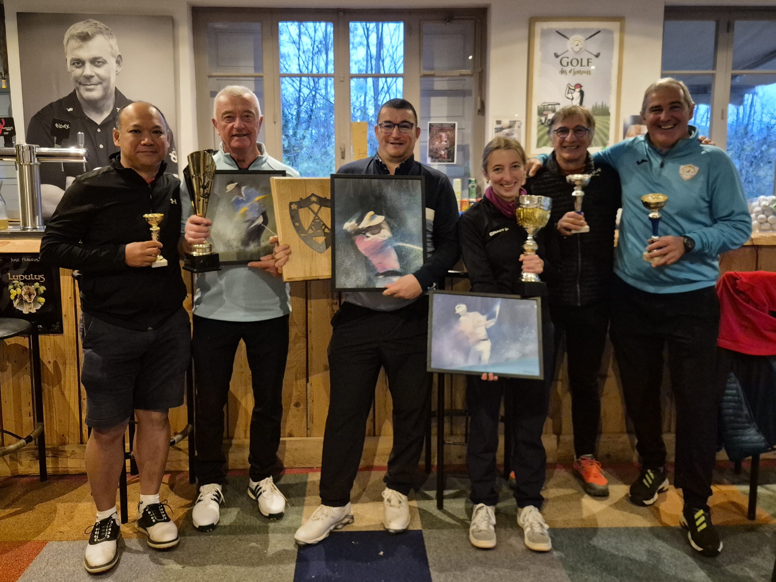 Groupe de golfeurs célébrant leurs succès lors d'un tournoi. Ils tiennent des trophées et des prix, entourés d'une ambiance conviviale dans un club de golf. En arrière-plan, des éléments de décoration liés au golf et des affiches. Les participants affichent des sourires, témoignant de l'esprit sportif et de camaraderie. Cette image illustre la passion pour le golf et l'importance des compétitions amicales.