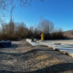 Groupe de personnes marchant sur un chemin en béton au bord d'un terrain dégagé, entouré d'arbres dénudés sous un ciel bleu clair. L'un des individus porte une combinaison jaune, tandis que d'autres sont vêtus de vêtements d'hiver. Ce paysage évoque une journée en plein air, propice aux activités de groupe et à la nature.