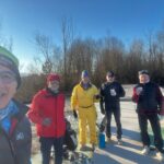 Groupe d'amis souriants sur un terrain de golf en hiver, profitant d'une journée ensoleillée. Ils portent des vêtements chauds, incluant des bonnets et des vestes colorées. Certains tiennent des gobelets, suggérant qu'ils savourent des boissons chaudes. En arrière-plan, un paysage naturel avec des arbres dénudés, typique de la saison froide. L'image évoque une ambiance conviviale et sportive, idéale pour les passionnés