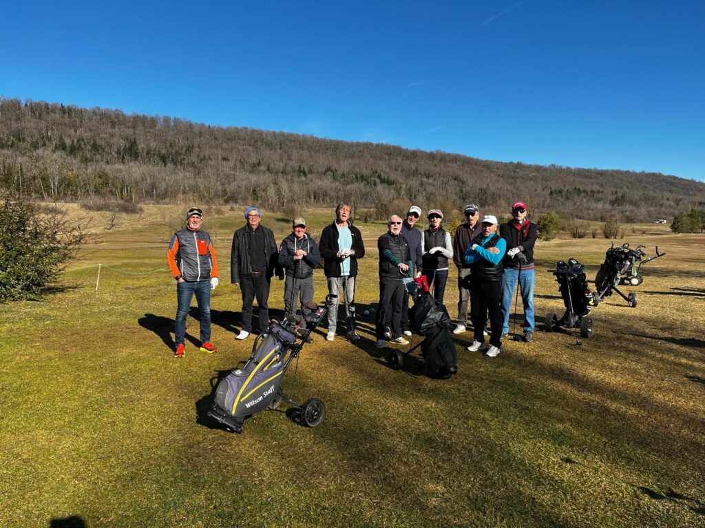 Groupe de golfeurs sur un terrain de golf par une journée ensoleillée. Les participants sont rassemblés sur le green, équipés de sacs de golf et prêts à jouer. En arrière-plan, un paysage naturel avec des collines et des arbres. L'image illustre une activité de loisirs en plein air, idéale pour les amateurs de golf.