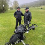 Deux golfeurs souriants se tiennent sur un parcours de golf sous une légère neige. Ils portent des vêtements imperméables, adaptés aux conditions météorologiques. À leurs côtés, une voiturette de golf équipée pour transporter des clubs et du matériel, témoignant de leur passion pour le golf même par temps frais. En arrière-plan, un paysage verdoyant et des arbres ajoutent à l'ambiance naturelle du lieu. Cette scène illustre la camaraderie et la détermination des gol