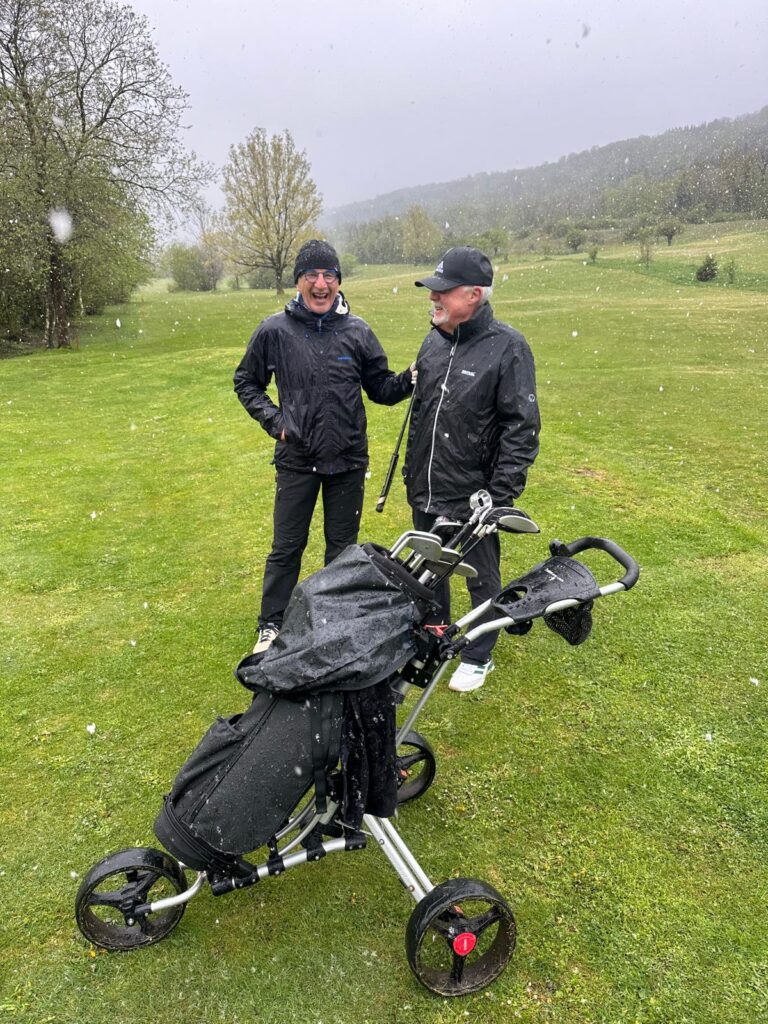 Deux golfeurs sur un terrain de golf par temps pluvieux, discutant près d'un chariot de golf. Ils portent des vêtements imperméables adaptés aux conditions météorologiques. L'arrière-plan montre un paysage verdoyant avec des arbres et des collines. Cette image illustre l'importance de la préparation pour jouer au golf, même sous la pluie.
