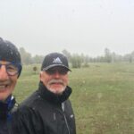 Deux golfeurs souriants sous la neige, vêtus de vêtements imperméables sur un parcours de golf verdoyant. La scène montre une ambiance hivernale inattendue, avec des flocons de neige tombant sur le terrain. L'arrière-plan présente un paysage de golf typique, avec des arbres et une pelouse bien entretenue. Cette image illustre la passion pour le golf, même par temps froid, et met en avant la camaraderie entre amis dans un cadre