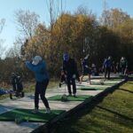 Groupe de golfeurs s'entraînant sur un practice en plein air, entouré d'arbres aux feuilles d'automne. Les participants, équipés de clubs de golf, travaillent leur swing sur des tapis verts. Des sacs de golf sont visibles à proximité, illustrant une ambiance conviviale et sportive. Cette scène met en avant l'importance de la pratique dans le golf, idéale pour les passionnés souhaitant améliorer leur technique.