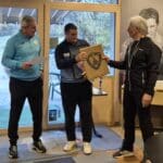 Cérémonie de remise de prix au sein d'un club sportif, mettant en scène trois hommes. L'un d'eux reçoit un trophée en bois, tandis que les autres l'observent avec intérêt. L'arrière-plan montre des éléments liés au sport, créant une atmosphère conviviale et festive. Cette image illustre l'esprit de compétition et de camaraderie dans le cadre d'un événement sportif.