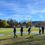 Un groupe de golfeurs s'entraîne sur un terrain de golf par une belle journée d'automne. Plusieurs joueurs sont alignés, concentrés sur leur coup, tandis que des flaques d'eau sont visibles sur le green, témoignant des récentes pluies. En arrière-plan, des arbres aux feuillages colorés et un ciel dégagé ajoutent à l'ambiance sereine de cette séance de golf.