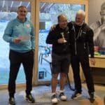 Célébration d'une remise de prix lors d'un événement sportif, avec trois hommes souriants. L'un des hommes tient un trophée, tandis que les autres l'applaudissent. L'arrière-plan montre des tables avec des récompenses et une décoration sobre. L'atmosphère est conviviale et festive, soulignant l'esprit d'équipe et la camaraderie au sein de la communauté sportive.