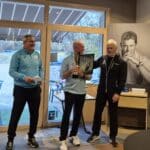 Cérémonie de remise de prix dans un club de golf, mettant en scène trois hommes souriants. L'un des hommes, au centre, tient un trophée et une récompense, tandis que les autres l'applaudissent. L'ambiance est chaleureuse et festive, avec des décorations de golf visibles en arrière-plan. Cette image illustre l'esprit sportif et la camaraderie au sein de la communauté golfique.