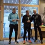 Cérémonie de remise de prix dans un club sportif, mettant en avant un gagnant tenant un trophée. Trois hommes sont présents, l'un lisant un discours, tandis que les autres célèbrent le lauréat. En arrière-plan, des affiches et des trophées sont visibles, créant une ambiance conviviale et festive. Cette image illustre l'esprit de compétition et de camaraderie au sein des activités sportives.
