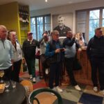 Groupe de personnes rassemblées dans un bar, apparemment après une activité de golf. Ils portent des vêtements de sport décontractés, certains tenant des verres. L'intérieur est décoré de bouteilles de vin et de trophées, avec une grande photo en noir et blanc accrochée au mur. L'ambiance semble conviviale et détendue, reflétant un moment de partage entre passionnés de golf.