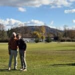 Deux golfeurs souriants posent sur un parcours de golf par une belle journée ensoleillée. L'arrière-plan présente des arbres aux feuillages d'automne et des collines, tandis que des chariots de golf sont visibles à proximité. L'image reflète une atmosphère conviviale et décontractée, idéale pour les amateurs de golf et les activités en plein air.