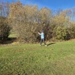 Un jeune golfeur s'apprête à frapper une balle sur un parcours de golf entouré d'arbres aux feuillages automnaux. Le ciel est dégagé et ensoleillé, créant une atmosphère agréable pour pratiquer ce sport en plein air. Des cônes de signalisation sont visibles sur le sol, indiquant la zone de jeu. Cette image illustre parfaitement l'enthousiasme et la concentration d'un amateur de golf lors d'une journée d'aut