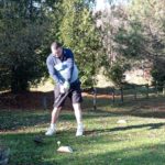 Homme en train de jouer au golf sur un terrain verdoyant, portant un pull à manches longues et un short. Il se prépare à frapper une balle de golf avec un club, entouré d'arbres et de feuilles au sol. L'image illustre une activité de plein air, idéale pour les amateurs de golf, mettant en avant la concentration et la technique du joueur.