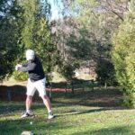Un golfeur en train de préparer son swing sur un parcours de golf verdoyant. Il porte un haut noir, un short blanc et une casquette blanche, se tenant sur une pelouse parsemée de feuilles. En arrière-plan, on aperçoit des arbres et une clôture en bois, créant une ambiance naturelle propice à la pratique du golf. Cette image illustre l'importance de la technique et de la concentration dans ce sport.