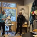 Deux hommes se tiennent dans un espace intérieur, probablement lors d'un événement lié au golf. L'un des hommes, vêtu d'un sweat bleu, lit un document tandis que l'autre, en tenue noire, sourit. Sur une table devant eux, plusieurs trophées, dont celui de la Ryder Cup 2018, sont exposés. À l'arrière-plan, des vêtements de golf et des accessoires sont accrochés sur un mur, ajoutant une ambiance sportive à la scène.