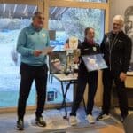 Cérémonie de remise de prix dans un club sportif, mettant en avant une jeune athlète tenant un trophée et une photo. Un homme, probablement un entraîneur, lit un discours de félicitations, tandis qu'un autre homme se tient à côté de la lauréate. L'ambiance est conviviale, avec des trophées exposés sur une table. En arrière-plan, on aperçoit des véhicules et la nature environnante, soulignant le cadre agréable de l'év