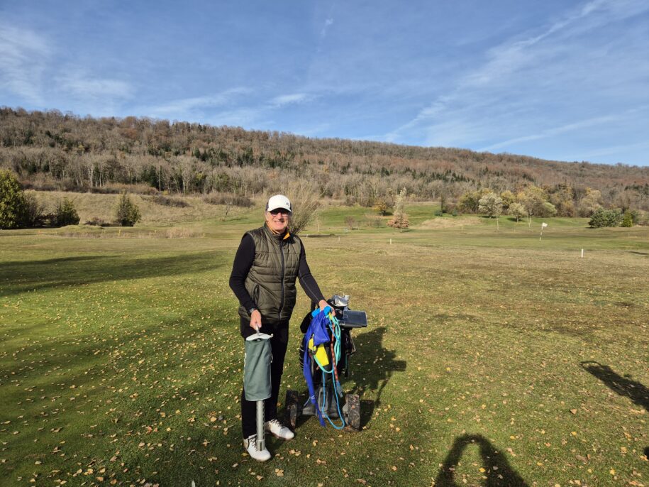 Un golfeur souriant se tient sur un parcours de golf verdoyant, entouré de collines boisées en arrière-plan. Il porte un gilet et une casquette, tenant un sac de golf rempli d'équipement. Le ciel est dégagé et ensoleillé, et le sol est parsemé de feuilles d'automne. Cette scène illustre une belle journée de golf, idéale pour les amateurs de ce sport en plein air.