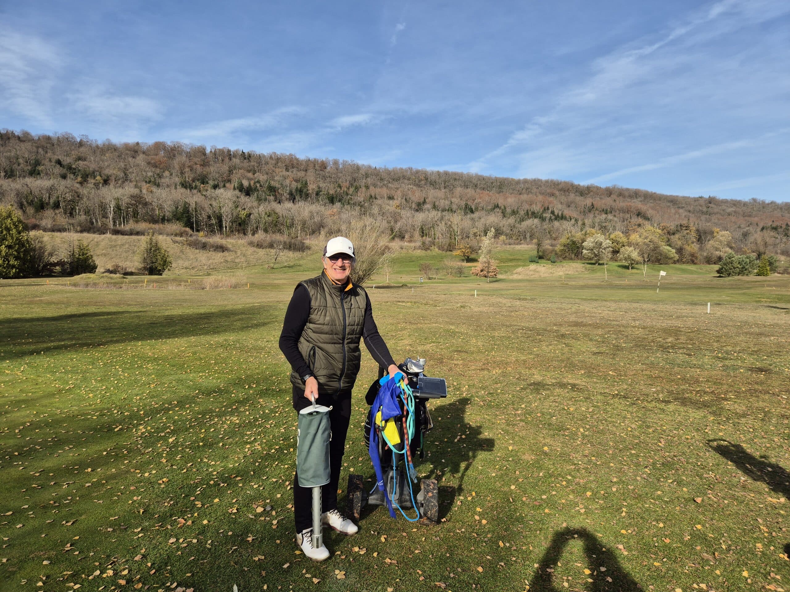 Un golfeur souriant se tient sur un parcours de golf verdoyant, entouré de collines boisées en arrière-plan. Il porte un gilet et une casquette, tenant un sac de golf rempli d'équipement. Le ciel est dégagé et ensoleillé, et le sol est parsemé de feuilles d'automne. Cette scène illustre une belle journée de golf, idéale pour les amateurs de ce sport en plein air.