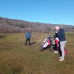 Deux golfeurs sur un terrain de golf par une journée ensoleillée. L'un des golfeurs, vêtu d'une veste bleu, se tient de dos, tandis que l'autre, portant un bonnet rouge, prépare son équipement à côté de ses sacs de golf. En arrière-plan, on aperçoit des arbres dénudés et un drapeau indiquant le trou. Cette scène illustre une sortie de golf, idéale pour les amateurs de ce sport en plein air.