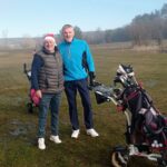 Deux golfeurs posent sur un terrain de golf par une journée ensoleillée, l'un portant un bonnet de Noël. Ils sourient et se tiennent près de leurs sacs de golf, équipés de clubs et de balles. En arrière-plan, on aperçoit un paysage verdoyant avec des arbres et un ciel clair, illustrant une atmosphère conviviale et sportive. Cette image évoque le plaisir du golf et les moments partagés entre amis.