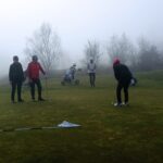 Groupe de golfeurs sur un green embrumé, pratiquant leur putting par une matinée brumeuse. Deux joueurs se tiennent debout, observant un autre golfeur en action, tandis qu'un quatrième membre du groupe attend avec un chariot de golf en arrière-plan. L'atmosphère calme et mystérieuse de la brume ajoute une dimension unique à cette séance de golf.