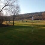 Vue panoramique d'un parcours de golf au crépuscule, mettant en avant un green avec un drapeau blanc. En arrière-plan, des collines boisées et des arbres dénudés créent une atmosphère paisible. Le sol est recouvert d'herbe verte bien entretenue, tandis que le ciel est dégagé, illuminé par la lumière douce du soleil couchant. Cette image évoque la tranquillité et la beauté d'une journée de golf en pleine nature.